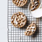 Close-up shot of nut-free cinnamon roll chaffles, sprinkled with cinnamon and monk fruit sweetener coating and drizzled with icing. The nut-free cinnamon roll chaffles are resting on a baking rack, atop a marble countertop.