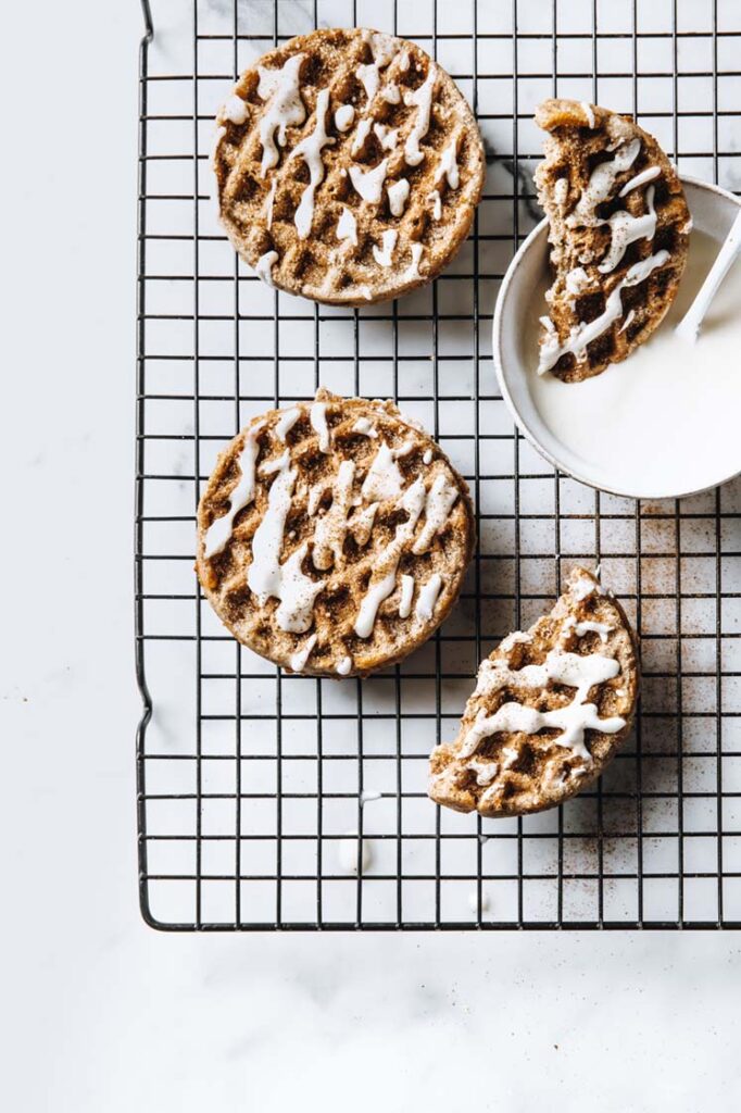 Close-up shot of nut-free cinnamon roll chaffles, sprinkled with cinnamon and monk fruit sweetener coating and drizzled with icing. The nut-free cinnamon roll chaffles are resting on a baking rack, atop a marble countertop.