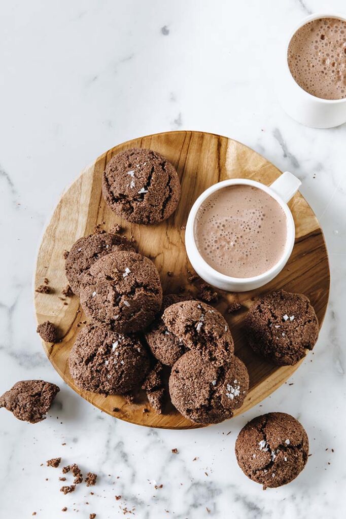 keto chocolate sea salt cookies on a wooden plate served with hot coco atop a marble kitchen table