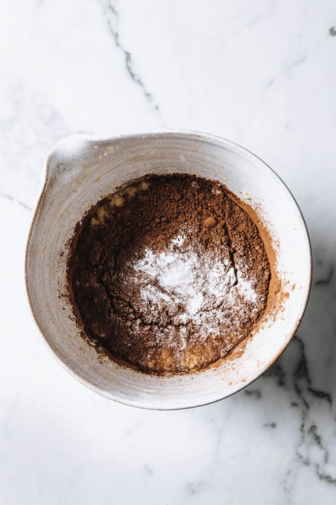monk fruit sweetener, raw cacao, baking powder and salt added on a mixture of coconut oil, eggs and vanilla extract in a bowl atop a marble kitchen table