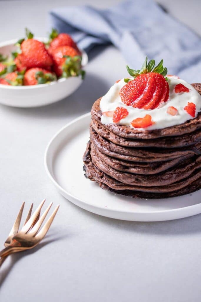 angled shot of chocolate protein pancakes on a plate topped with strawberries and cream