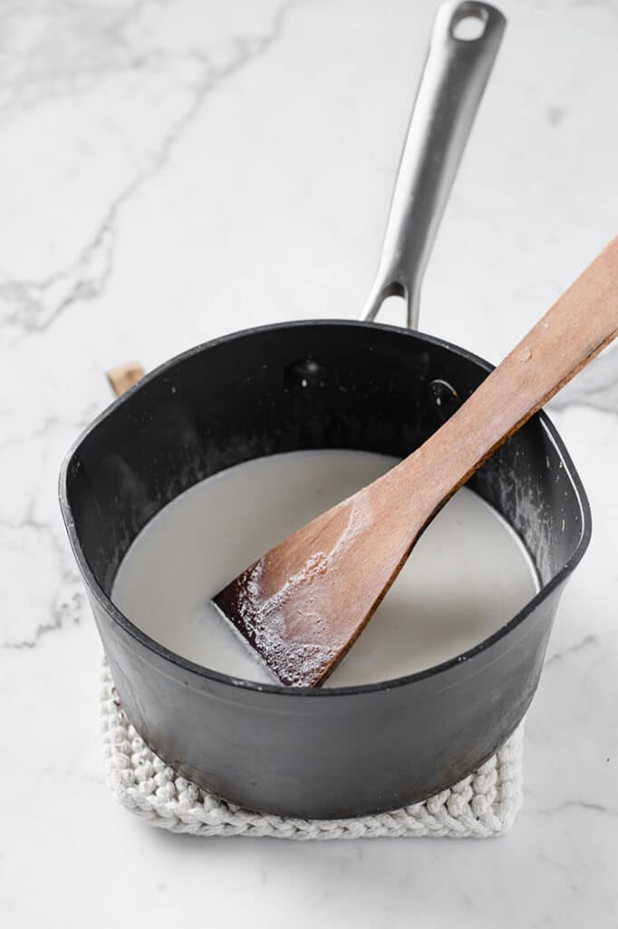 a pot of coconut milk and bloomed gelatin being stirred with wooden spatula atop a marble kitchen counter
