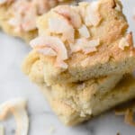 Close-up shot of stacks of low-carb coconut blondies and scattered coconut flakes, atop a marble countertop.