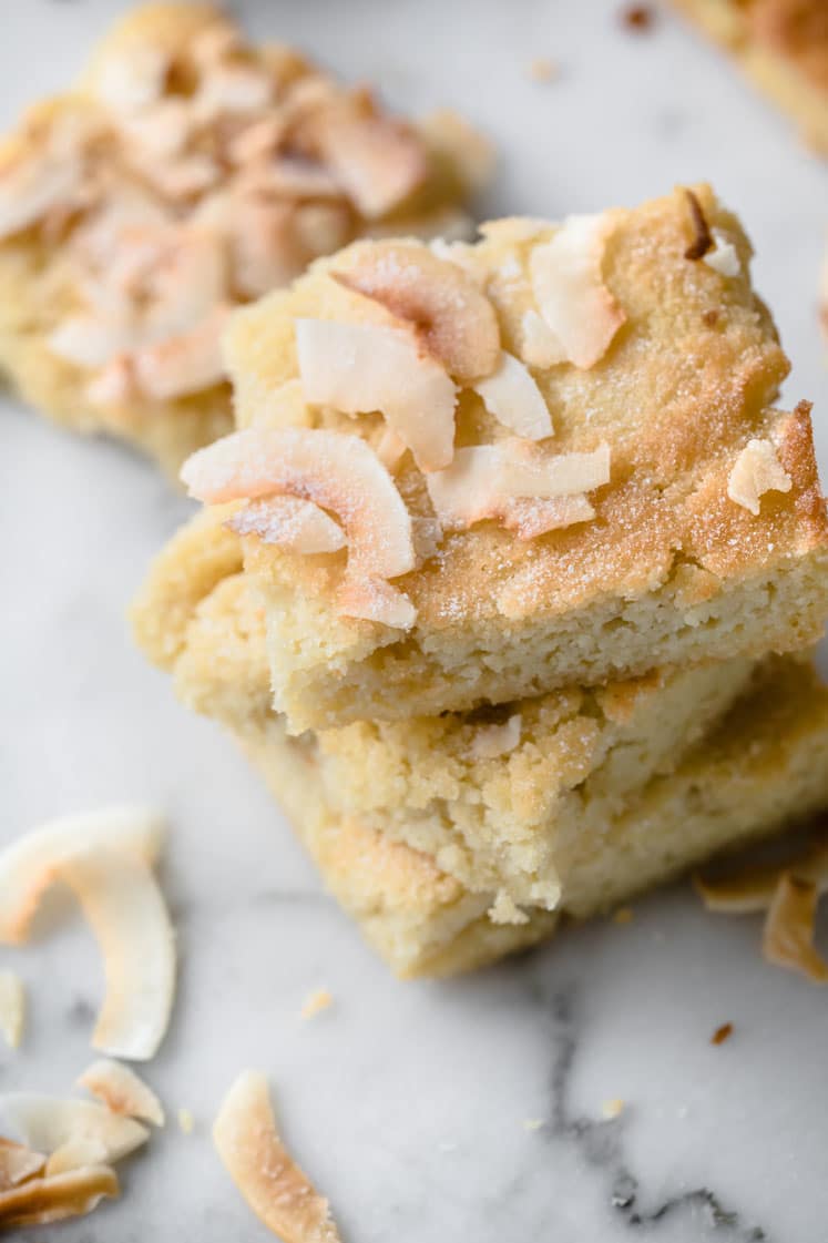 Close-up shot of stacks of low-carb coconut blondies and scattered coconut flakes, atop a marble countertop.