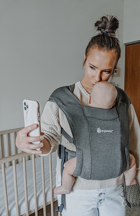 mother carrying baby using ergobaby carrier while taking a photo with cellphone