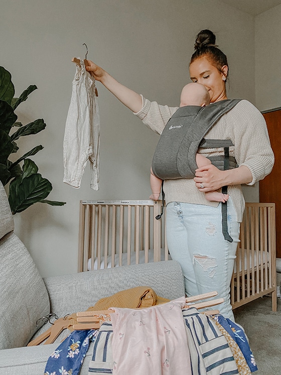 mom holding up outfit while looking down at baby in ergobaby carrier in nursery