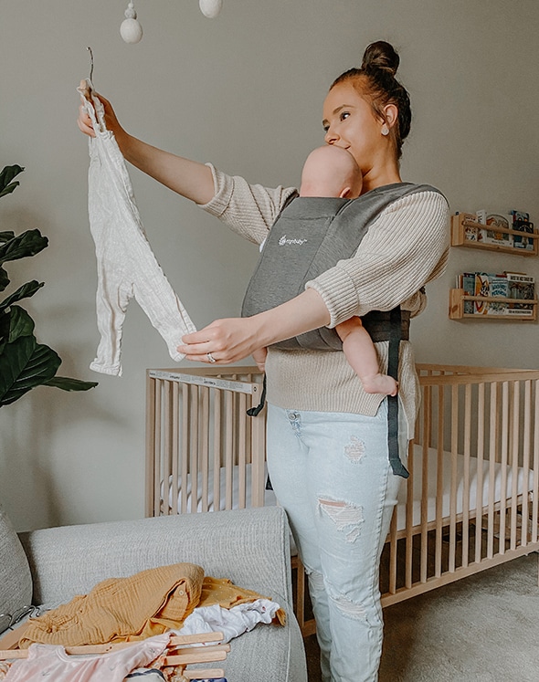 mom looking at baby outfit while holding baby in baby carrier in nursery