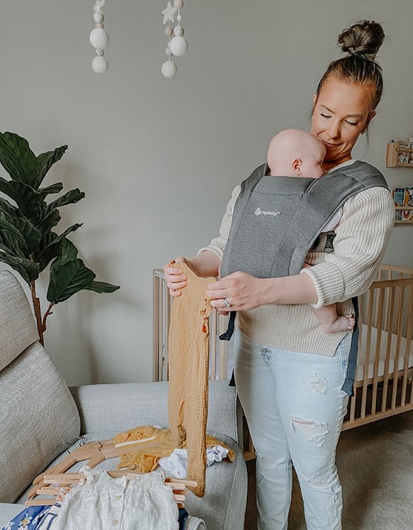 mom holding baby in ergobaby embrace carrier while folding baby clothes