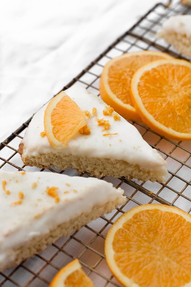 Angled, close-up shot of low-carb glazed orange scones and a few orange slices resting on a baking rack, atop a marble countertop.