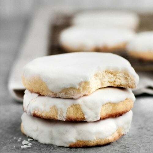 Close-up shot of a stack of low-carb glazed sugar cookies atop a marble countertop. In the background, there’s a baking pan with a few low-carb glazed sugar cookies.