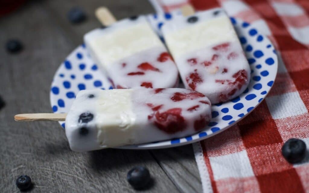 4th of July Keto Recipes - 3 red,white, & blue keto popsicles on a blue polka dotted plate atop a wooden table