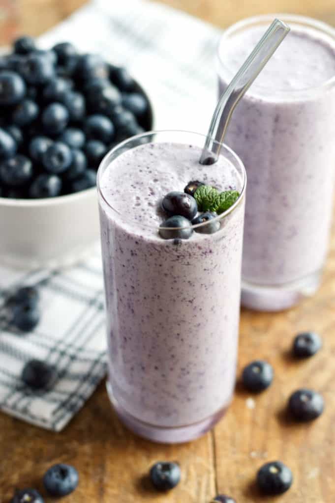a glass of blueberry keto smoothie with a bowl of blueberries on the background atop a wooden table