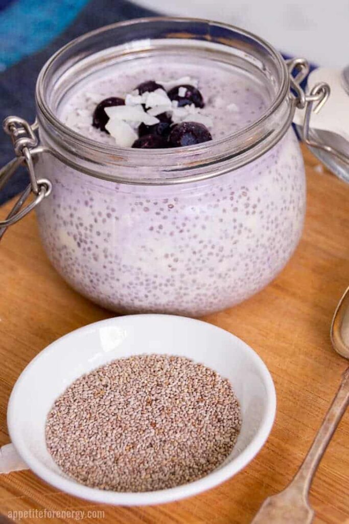 keto berry recipes: angled view of a bowl of chia seeds with a glass jar of low carb blueberry chia pudding behind atop a wooden table