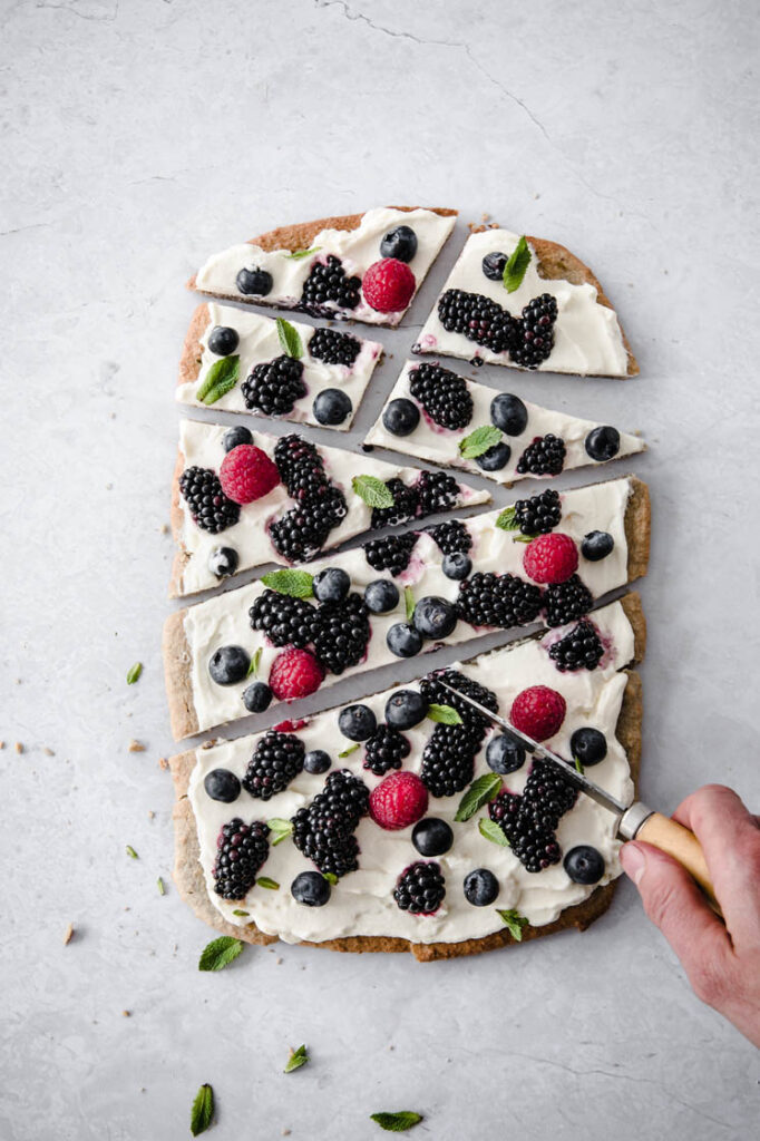 overhead image of keto fruit pizza being sliced to irregular pieces atop a marble kitchen counter