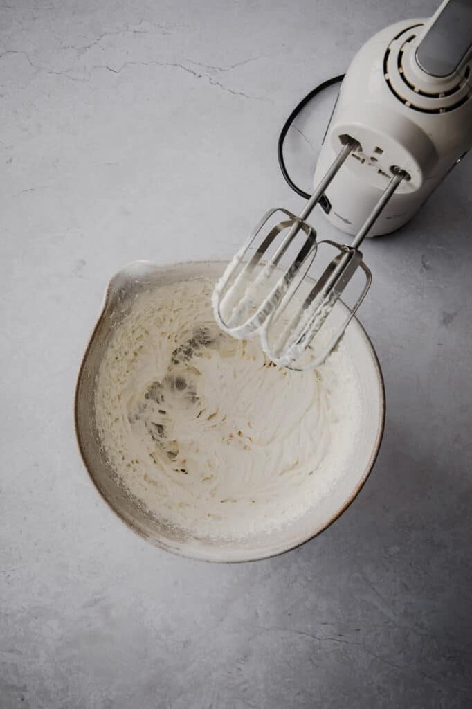 keto fruit pizza cream cheese frosting being mixed with an electric mixer in a large mixing bowl atop a marble kitchen table.