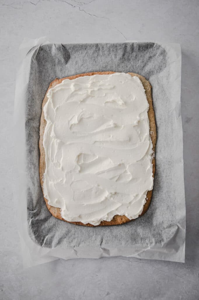 cream cheese frosting spread on top of crust on a baking sheet lined with parchment paper atop a marble kitchen counter.