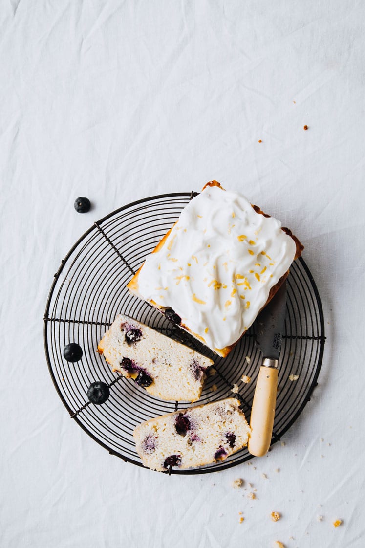 a loaf of keto lemon blueberry bread with greek yogurt frosting resting on a wire rack atop a marble kitchen table