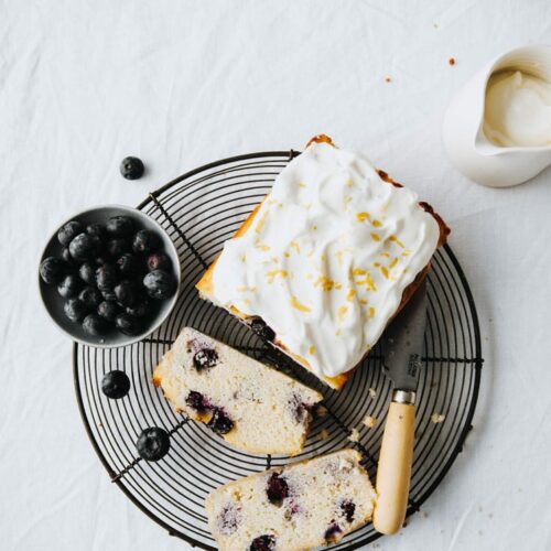 a loaf of keto lemon blueberry bread with greek yogurt frosting resting on a wire rack with a bowl of blueberries atop a marble kitchen table