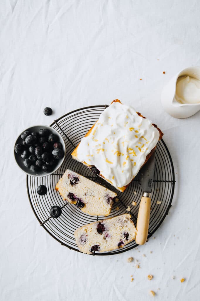 a loaf of keto blueberry bread with greek yogurt frosting resting on a wire rack with a bowl of blueberries atop a marble kitchen table