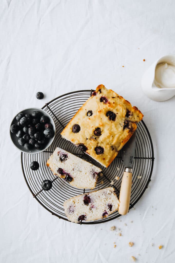 keto lemon bread topped with blueberries resting on a wire rack with a bowl of blueberries atop a marble kitchen table