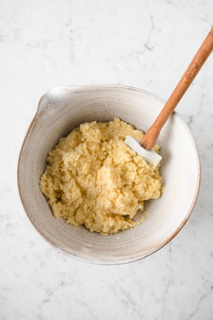 keto lemon blueberry dough in a mixing bowl being mixed by a rubber spatula atop a marble kitchen table