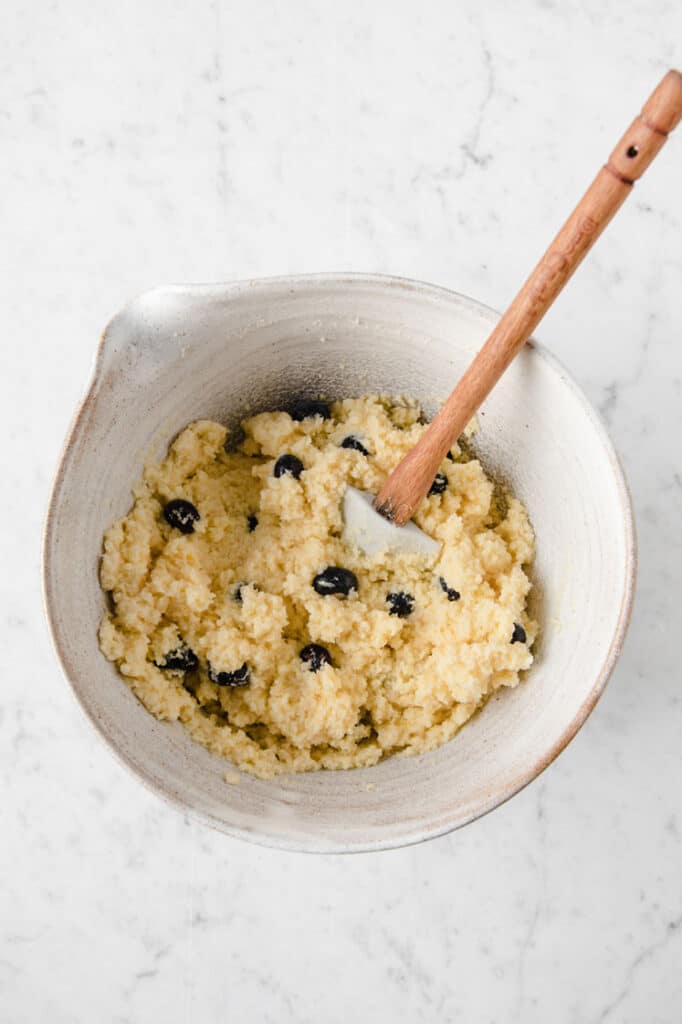 bread dough in a mixing bowl being mixed with blueberries by a rubber spatula atop a marble kitchen table