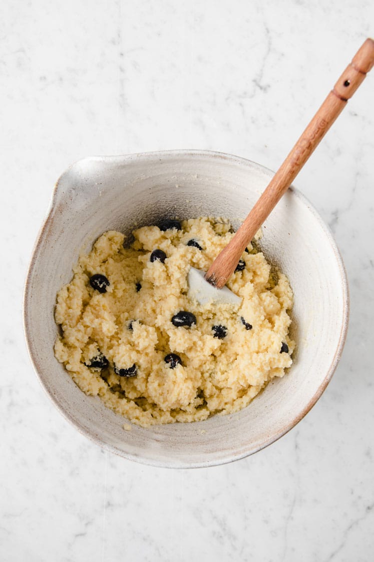 keto lemon blueberry dough in a mixing bowl being mixed with blueberries by a rubber spatula atop a marble kitchen table