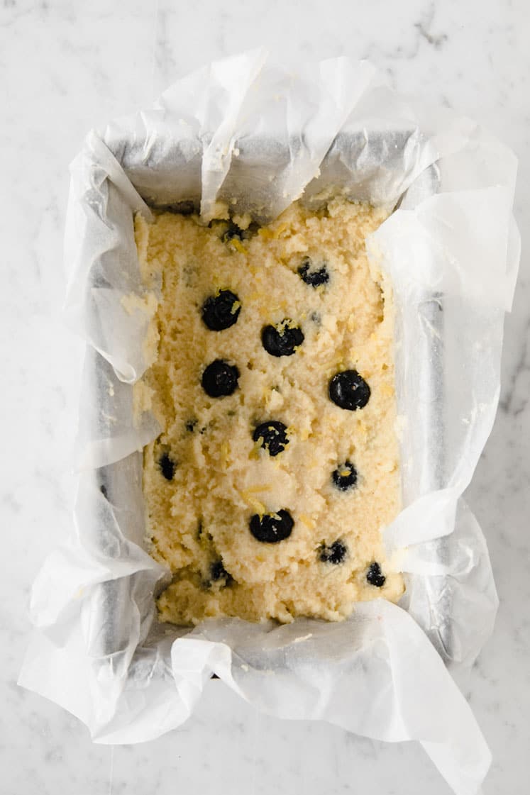 keto lemon blueberry bread dough in a loaf pan lined with parchment paper sprayed with non-stick cooking spray atop a marble kitchen table