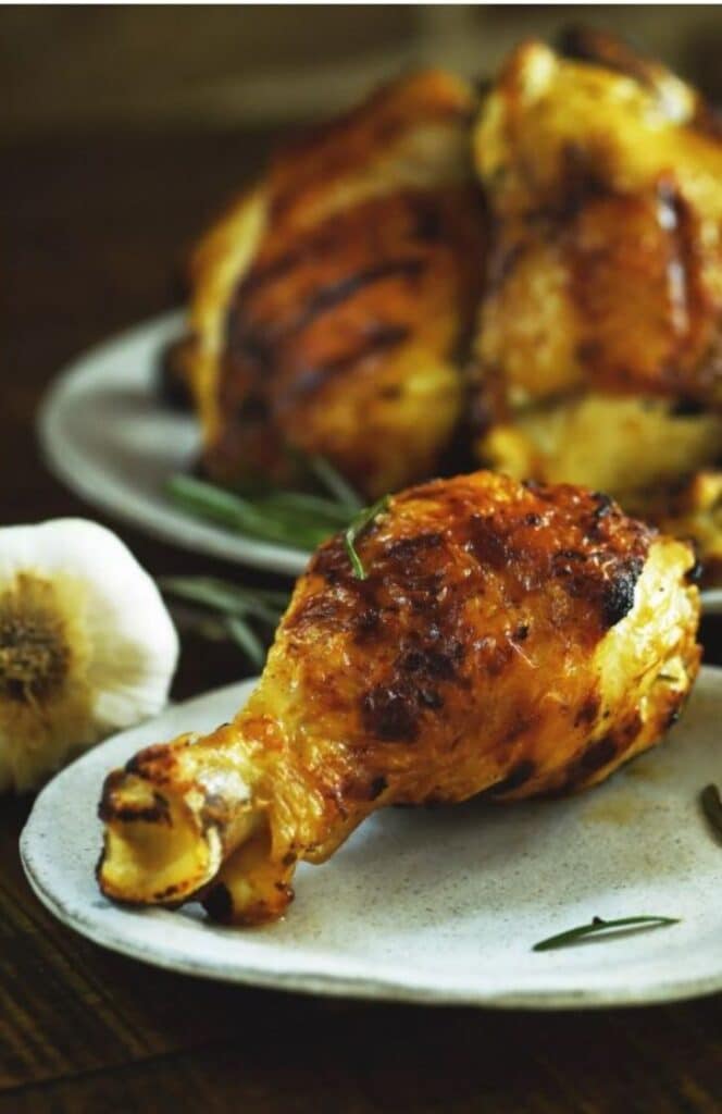 close-up image of grilled rosemary chicken drumstick on a blue plate atop a wooden table with the whole chicken on the background