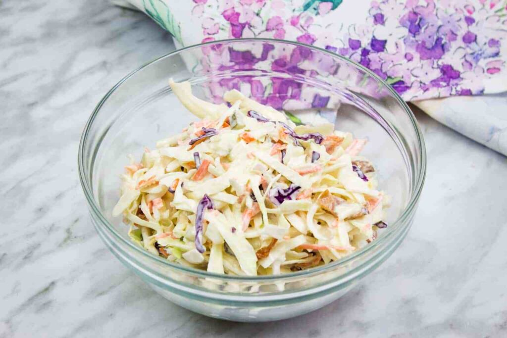 a clewa glass bowl of pickle slaw with bacon atop a marble kitchen table