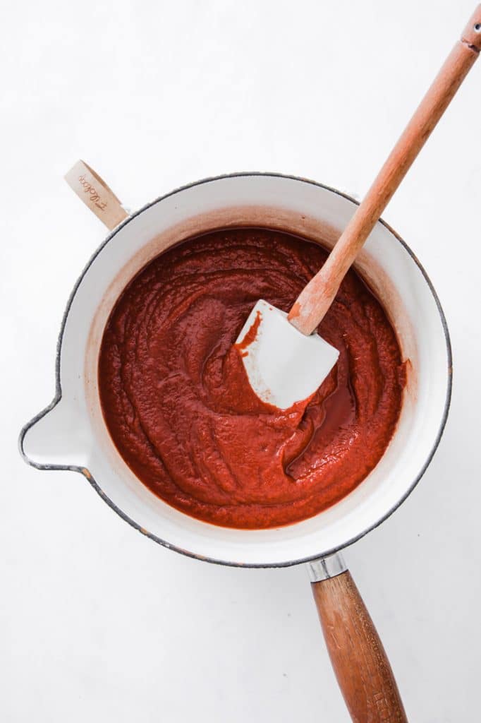 ketchup mixture being stirred with a rubber spatula in a medium-sized pan atop a marble kitchen counter
