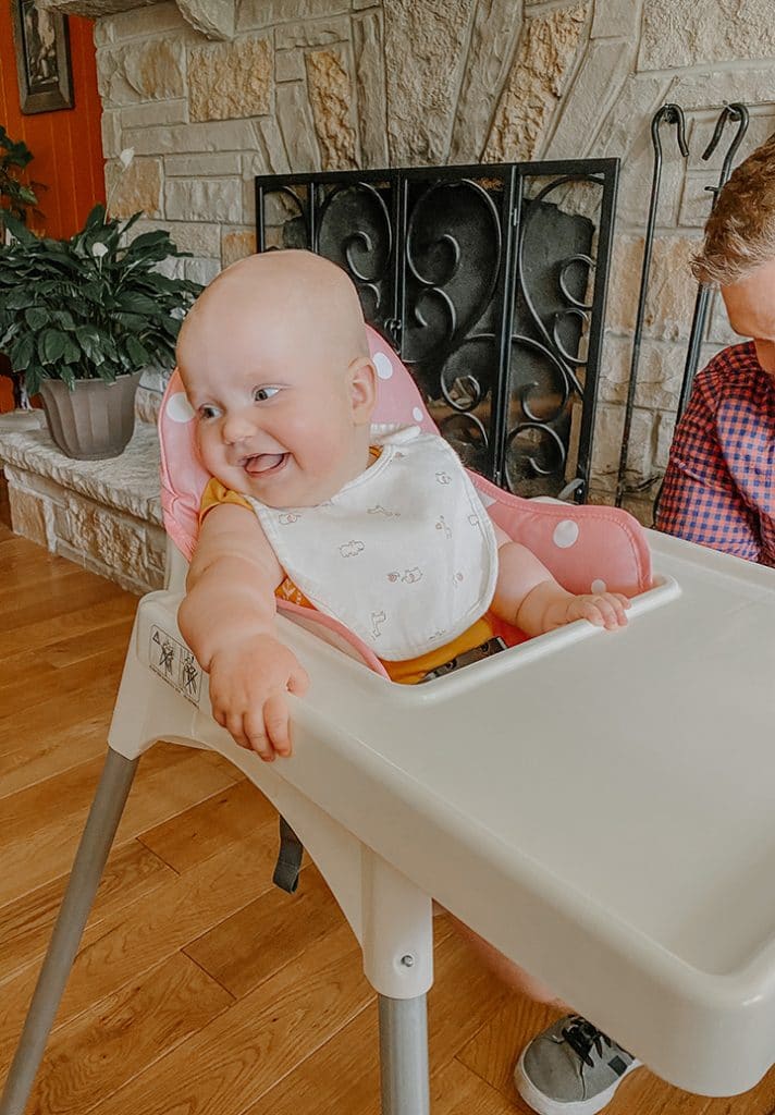 introducing baby to peanut butter while baby sits in a high chair looking to the side and smiling in front of fire place