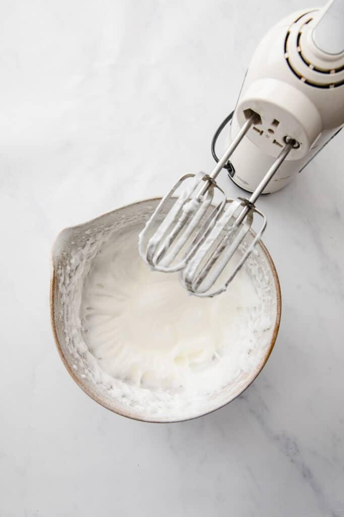 overhead shot of mixed egg whites in a mixing bowl with an electric mixer on the side atop a marble kitchen table