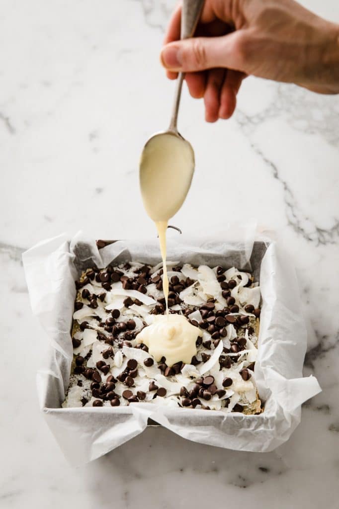 sweetened condensed milk being poured on top of a baking pan of keto magic bars