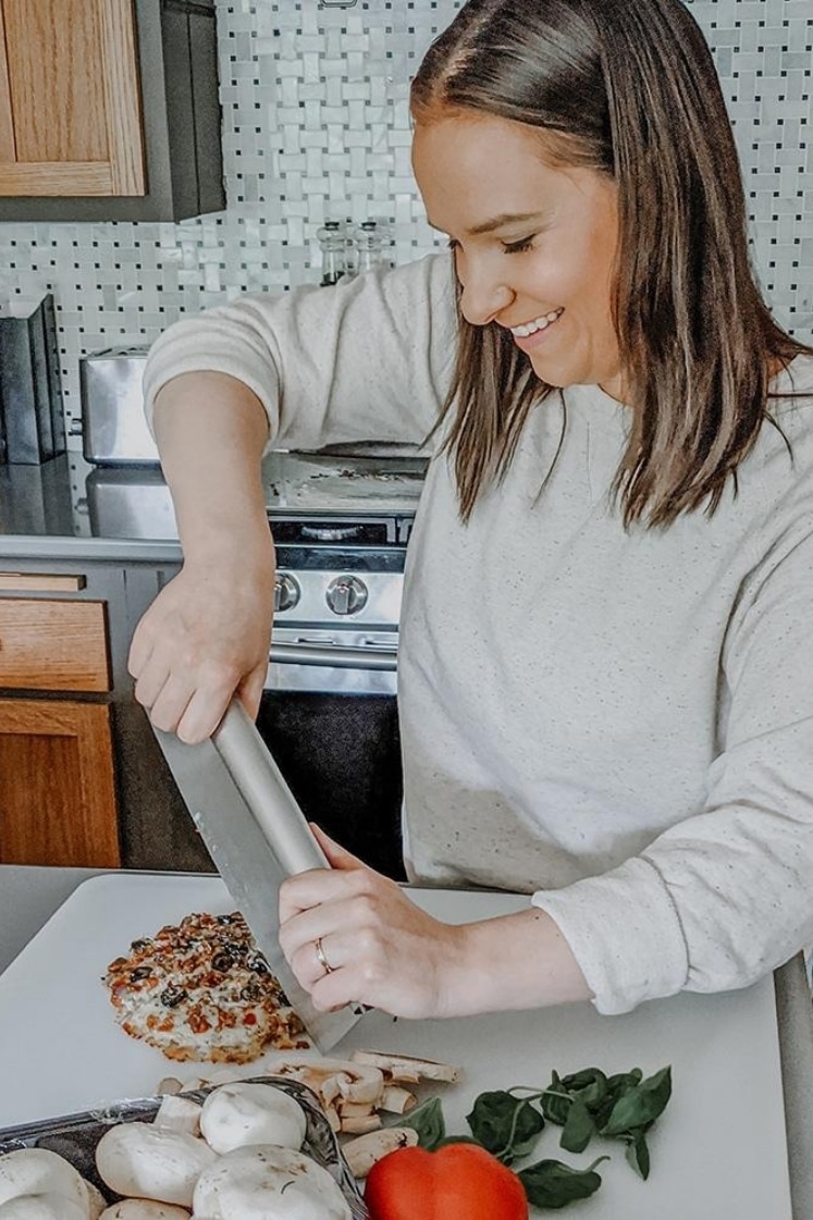 sara nelson cutting cooked frozen pizza with pizza cutter on a cutting board in a kitchen