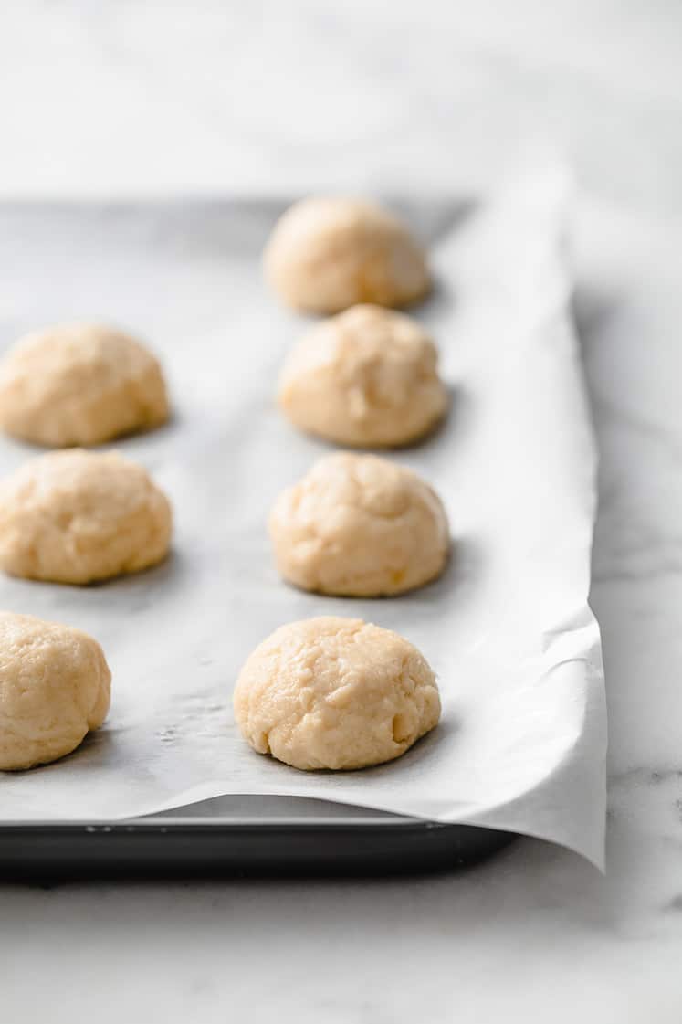 dough balls lined up on a baking pan lined with parchment paper atop a marble kitchen counter