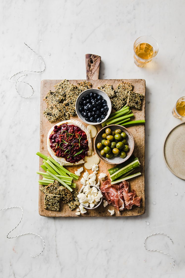 keto baked brie served on a serving board with nut-free crackers, celery, and a bowl of blue berries and olives atop a marble kitchen counter