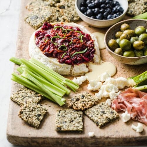 angled shot of baked brie with nut-free crackers, celery, a bowl of olives, and a bowl of blue berries on a serving board atop a marble kitchen counter