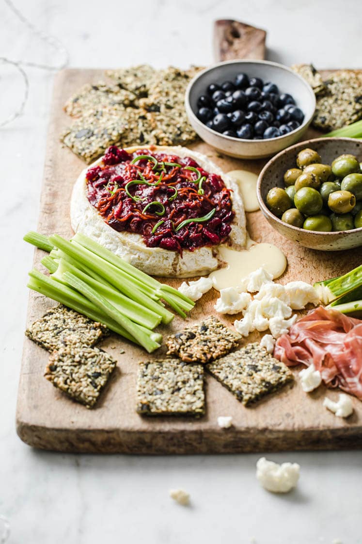 angled shot of baked brie with nut-free crackers, celery, a bowl of olives, and a bowl of blue berries on a serving board atop a marble kitchen counter