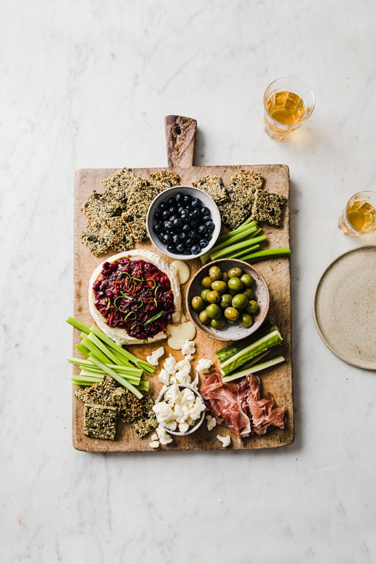overhead shot of baked brie with nut-free crackers, celery, a bowl of olives, and a bowl of blue berries on a serving board atop a marble kitchen counter