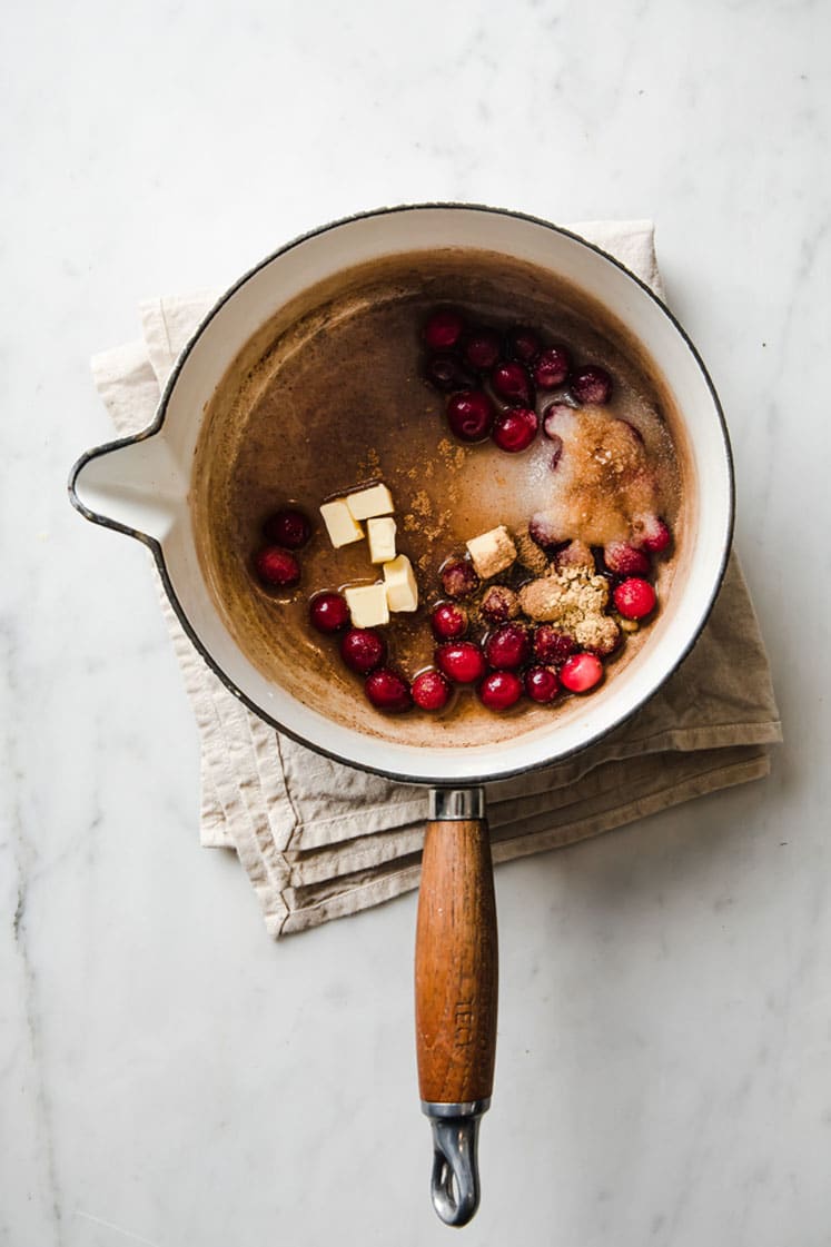 overhead image of butter, cranberries, , monk fruit sweetener, cinnamon, cardamom pods, nutmeg, ginger, and water in a sauce pan atop a marble kitchen counter