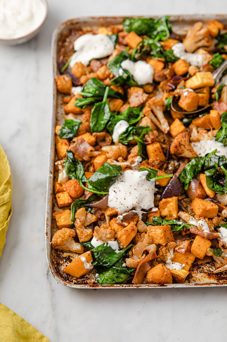 an angled image of a baking tray of roasted butternut squash and cauliflower and bacon topped with sour cream, a rolled yellow napkin and a bowl of sour cream on the left hand corner atop a marble kitchen counter