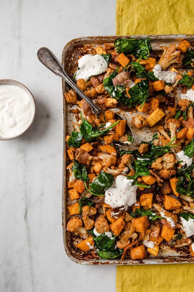 a baking tray of roasted butternut squash and cauliflower and bacon topped with sour cream on a yellow napkin with a bowl of sour cream on the left hand corner atop a marble kitchen counter
