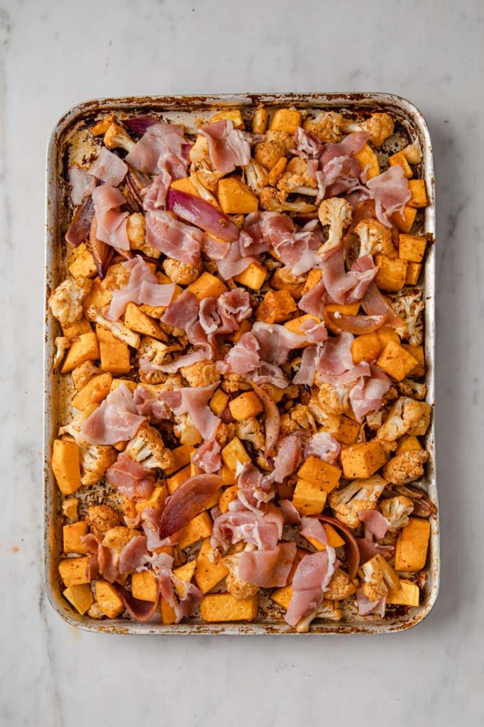 overhead image of butternut squash, cauliflower, and onion, topped with bacon on a baking tray atop a marble kitchen counter