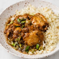 close-up image of a bowl of cauliflower rice topped with chicken adobo atop a marble kitchen counter
