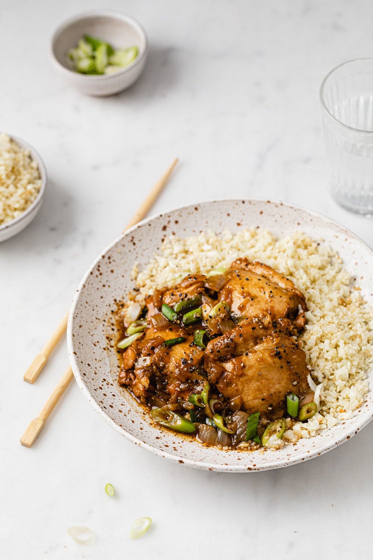 a bowl of cauliflower rice topped with chicken adobo with a pair of chopsticks place on the left sie atop a marble kitchen counter
