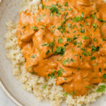 Overhead shot of a plate with cauliflower rice, topped with the Instant Pot Butter Chicken, garnished with chopped fresh parsley with a spoon, atop a marble kitchen counter.