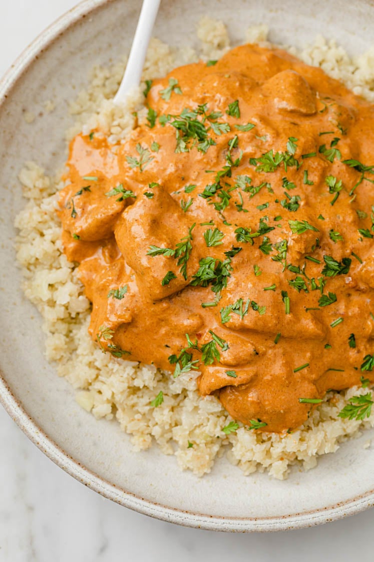 Overhead shot of a plate with cauliflower rice, topped with the Instant Pot Butter Chicken, garnished with chopped fresh parsley with a spoon, atop a marble kitchen counter.