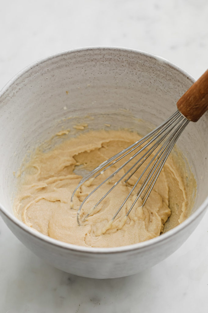 a mixture of monk fruit sweetener, cream of tartar, heavy cream and vanilla extract being whisked in a mixing bowl, atop a marble kitchen table.
