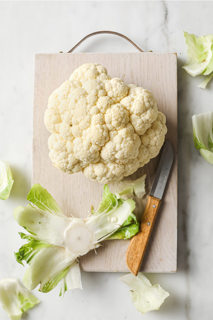 Overhead shot of cauliflower head with the bottom and leaves removed. Photographed on a wooden board with a small knife atop a marble countertop